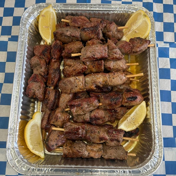 Grilled meat skewers with lemon wedges in a foil tray on a blue and white checkered tablecloth.