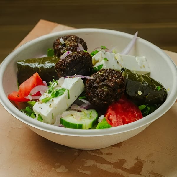 A Greek salad in a white bowl with falafel, feta, dolmades, tomatoes, cucumbers, and red onions.