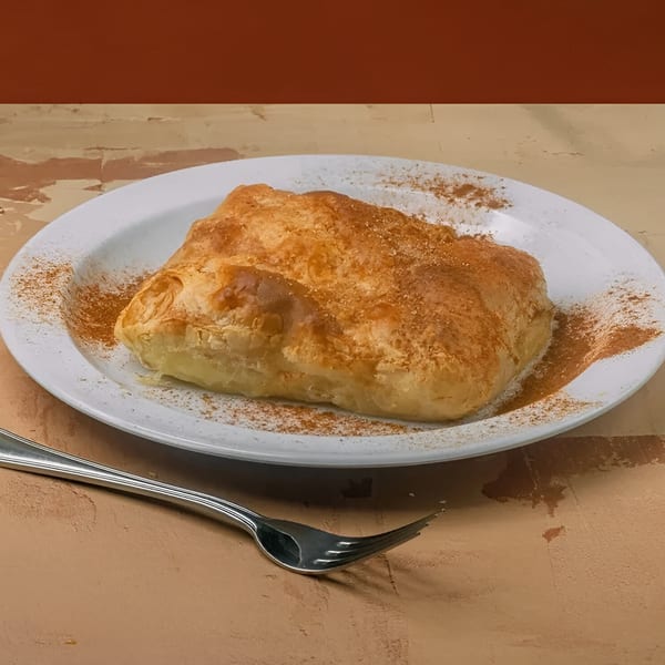 A golden-brown Greek bougatsa pastry, dusted with cinnamon, on a white plate with a fork.