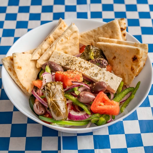 A Greek salad with feta, olives, tomatoes, onions, peppers, and pita bread on a checkered tablecloth.