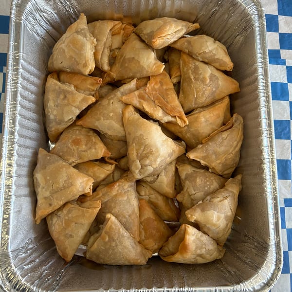 A foil tray filled with many golden-brown, triangular samosas on a blue and white checkered tablecloth.