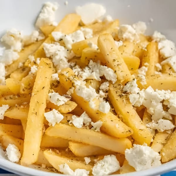 Close-up of golden Greek fries topped with crumbled feta cheese and dried herbs in a white bowl.