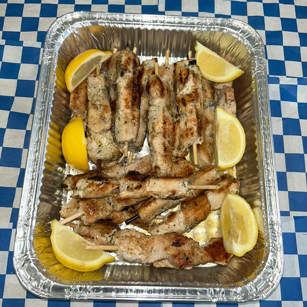 Grilled chicken souvlaki skewers with lemon wedges in a foil pan on a blue and white checkered tablecloth.