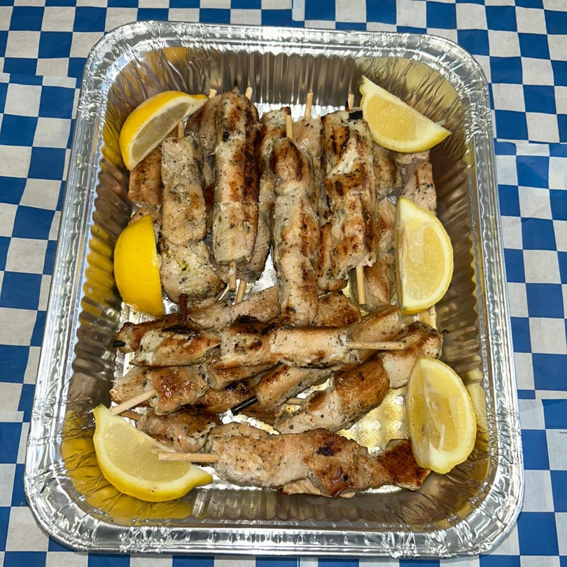 Grilled chicken souvlaki skewers with lemon wedges in a foil pan on a blue and white checkered tablecloth.