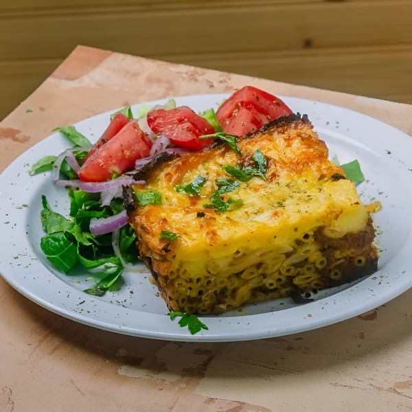 A golden-brown pastitsio with a side salad of tomatoes, red onion, and greens on a white plate.