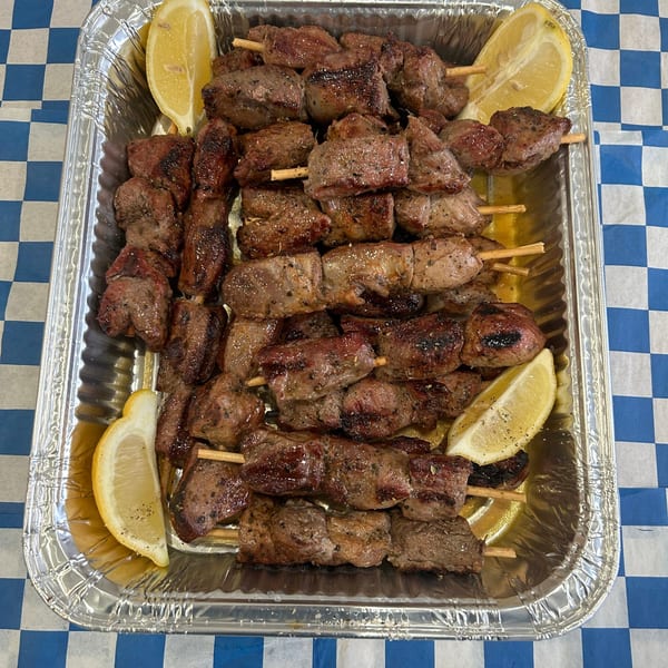 Grilled meat skewers with lemon wedges in a foil tray on a blue and white checkered tablecloth.